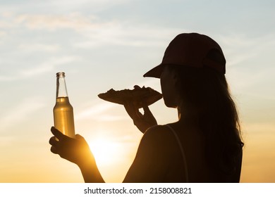 Silhouette Of Girl In Baseball Cap Eating Pizza And Drinking Soda Water From Glass Bottle And Looking At Sunset Sky. Rear View Of Female 