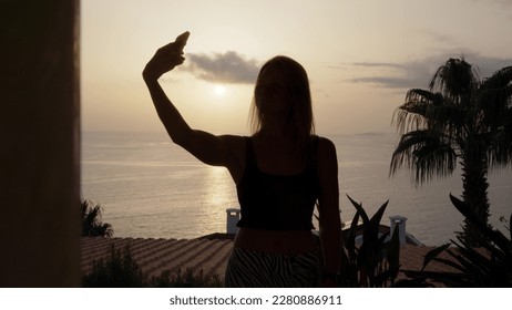 silhouette of girl in black top takes selfie on her phone against background of sunset or dawn on balcony of her room hotel. young woman takes pictures of herself on smartphone. - Powered by Shutterstock