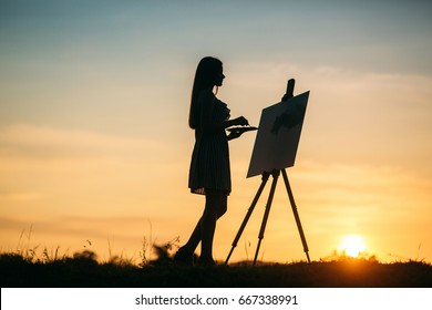 Silhouette of a girl. The blonde girl paints a painting on the canvas with the help of paints.