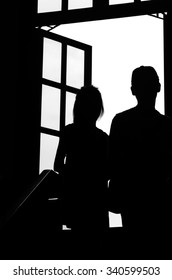 Silhouette Of A Girl And A Boy Walking Up Stairs. A Big Window In Front Of Them. Black And White Image.