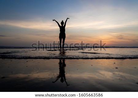 Silhouette of a girl standing in the water with her arms raised and her reflection in the water as the sun goes down