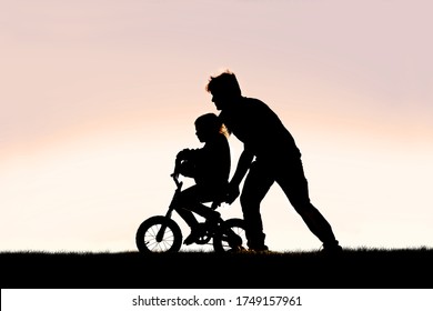 A Silhouette of a good father is helping his young girl child learn to ride her bicycle with training wheels on a summer day.