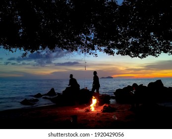 Silhouette of group of man with campfire at the rocky beach during sunset. Long exposure created blur sky background.
