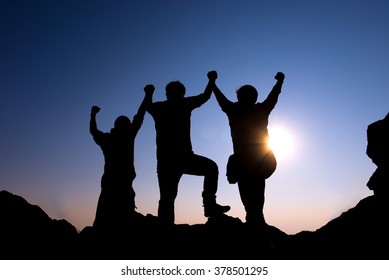 Silhouette Of Group People On The Peak Of Rocks Mountain At Sunset With Arms Raised Up Above Head In Celebration