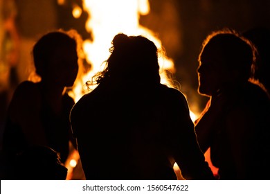 silhouette of group of people sitting in the front of the fire as they talk, seen from behind during dark night campfire, blurred background