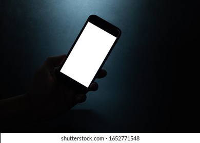 silhouette hand holding a mobile phone with blank wihte screen in the dark darkness against dark blue background  