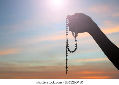 Silhouette of a hand holding Rosary on colorful sunset sky light background. Person with Rosary in hands with Jesus Christ crucifix. Copy space for spiritual text message design. Catholic symbol.