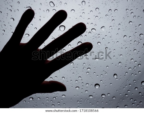 Silhouette of hand on glass with water drops in\
lonely rainy day on dim\
sky.