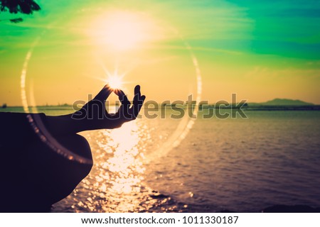 Silhouette, a hand of woman meditating in a Yoga pose or Lotus position at the Sea in Sunset and reflection of sunlight. Nature meditation and spa like zen or calm and solitude concept. Relax time.
