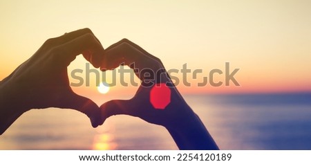 Silhouette of hands in the shape of a heart of love. Women's hands in the shape of a heart against the background of the sea and the setting sun let the sun's rays through, toned. Romantic background	