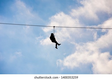 Silhouette Of A Hanging Man On A Zip Line In Front Of A Cloudy Blue Sky Background