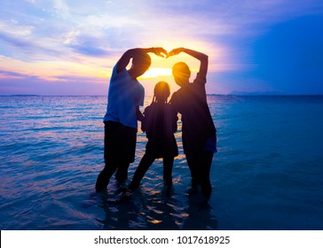 Silhouette der glücklichen asiatischen Familie in den Urlaub verliebt und glückliche Valentinstag. Papa und Mama, die Herzform Schild vorbei an Sonnenstrahlen stehen auf tropischem Strand mit Sonnenuntergang Himmel Hintergrund. Familienkonzept.