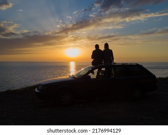 Silhouette of a happy couple sitting on the roof of a car on a cliff in front of the sea with a beautiful orange-blue sky at sunset, view from a drone. Concept of vacations and travel, romance - Powered by Shutterstock