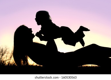 A Silhouette Of A Happy, Laughing Mom Is Laying In The Grass At Sunset, Lifting Her Baby Boy Up Into The Air Over Her Head