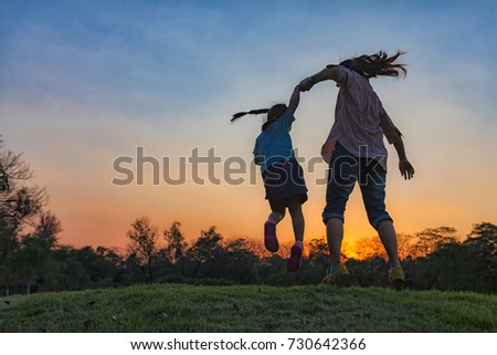Silhouette of a happy little girl child is jumping with his mother against the sunset on a summer day on Chatuchak Park at Bangkok City, Thailand. Concept of friendly family.