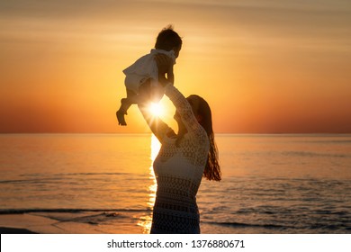 Silhouette Of A Happy Mother Holding Her Baby Up In The Air On A Beach During Sunset Time