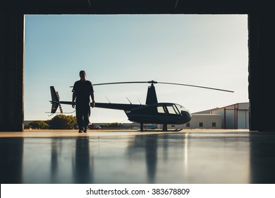 Silhouette Of Helicopter With A Pilot In The Airplane Hangar. Pilot Walking Away From Helicopter Parked Outside The Hangar.