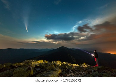 Silhouette of a hiker man with flashlight standing on mountain peak under starry sky lighting a beam og light on Neowise comet with light tail.