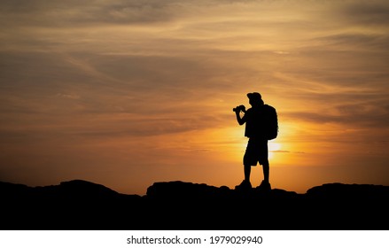 Silhouette Of Hiker Man Holding Binoculars On Sunset From Mountain Top. Old Man With Backpack Watching The Sunset