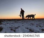  Silhouette of a Hunter holding a pheasant with a Retriever Dog at Sunset on a Snow-Covered Field 