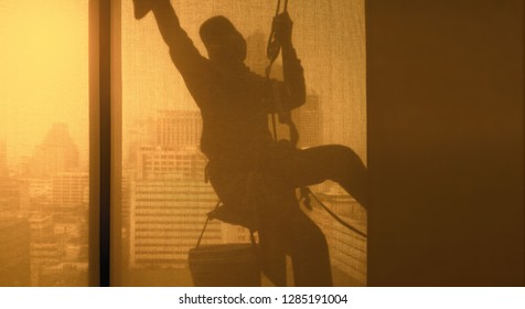 Silhouette Images And Shoot Through Transparent Curtain Effect From Inside Building Which A Man Cleaning The Window Of High Office Building With His Equipment Such As Wipe, Sponge, Bucket