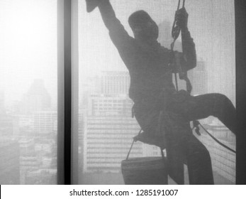 Silhouette Images And Shoot Through Transparent Curtain Effect From Inside Building Which A Man Cleaning The Window Of High Office Building With His Equipment Such As Wipe, Sponge, Bucket