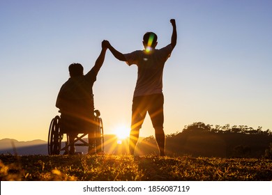 Silhouette Of Joyful Disabled Man In Wheelchair Raised Hands With Friend At Sunset