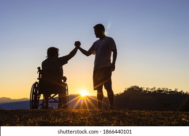 Silhouette of joyful disabled man in wheelchair raised hands with friend at sunset - Shutterstock ID 1866440101