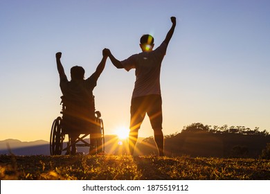 Silhouette Of Joyful Disabled Man In Wheelchair Raised Hands With Friend At Sunset