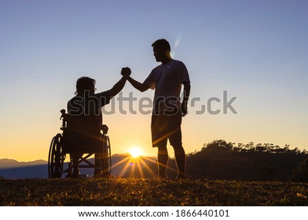 Silhouette of joyful disabled man in wheelchair raised hands with friend at sunset