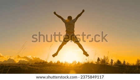 SILHOUETTE, LENS FLARE: Victory jump of a young guy on a mountaintop at sunrise. He is celebrating as he reaches the top when first rays of autumn morning sun start spilling over the alpine hilltop.