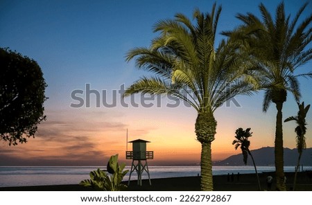 Silhouette of lifeguard tower and lush palm trees in the sunset view of the Mediterranean beach of Cala del Moral in Rincon de la Victoria, Malaga, Spain.