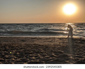 The silhouette of a little girl playing on the shore under the golden rays of the setting sun. The sea waves gently touch the shore, creating a magical atmosphere of serenity and childlike joy at dusk