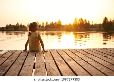 Silhouette of a little girl sitting on the pier and watching sunset by the lake. The beautiful light of the sunset is reflected in the water of the lake. - Powered by Shutterstock