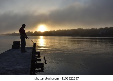 A  silhouette of a lone fisherman on a wooden dock. The sun behind is rising or setting in a gray, misty and ominous sky. The water is dark and calm. The man is facing right, on the left of photo. - Powered by Shutterstock