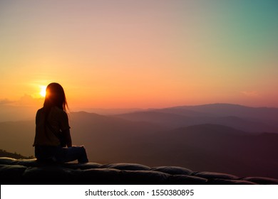 The silhouette of a lonely girl sitting on a bench watching the sunset on the top of the hill
