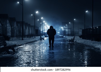 Silhouette of lonely person walks on dark foggy street illuminated with street lamps, blue toned