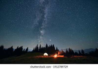 Silhouette Of Male Traveler Near Illuminated Camp Tent Under Beautiful Night Sky With Stars. Magnificent View Of Blue Starry Sky Under Grassy Hill. Concept Of Travelling, Hiking And Night Camping.