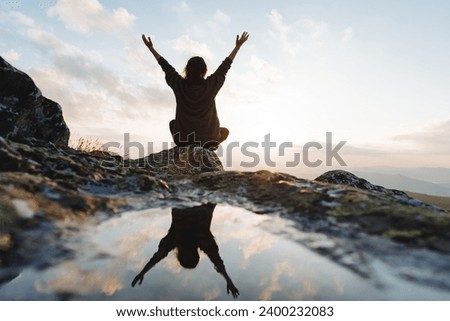 Silhouette of a man from the back sitting on a rock in the mountains, a joyful girl view from behind raised her hands up, reflection in a puddle of water on a stone, dawn in the mountains