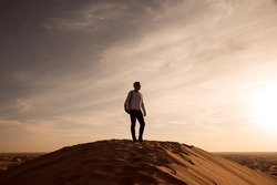 
Silhouette Of A Man With A Backpack Standing On A Dune At Sunset.