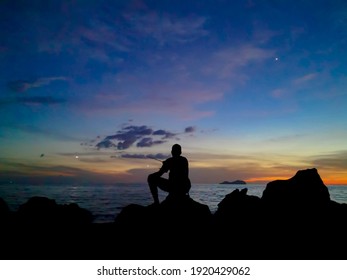 Silhouette of a man fishing and sitting on the rocks looking at the ocean during sunset. Long exposure creating motion blur of the background.