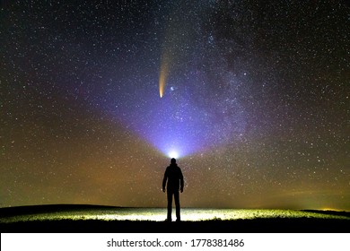 Silhouette of a man with flashlight on his head pointing bright beam of light on starry sky with Neowise comet with light tail.