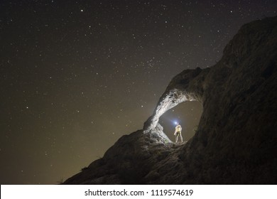 Silhouette Of Man With Flashlight Standing Under The Stone Arch In A Mountain Valley With Many Stars In A Night Sky.