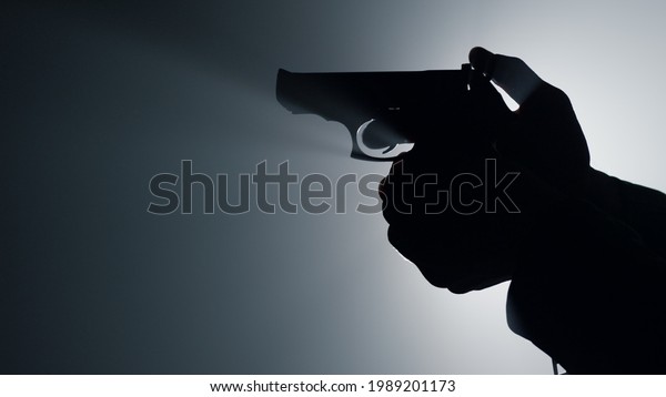 Silhouette of man hand holding\
gun indoors. Closeup unknown criminal arm cocking trigger in\
spotlight background. Unrecognizable male person shooting firearm\
in dark. 
