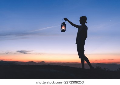 Silhouette of a man holding a lit kerosene lamp to light his way in the dark, with sky after sunset in the background. 
