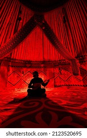 Silhouette Of Man Holding And Playing Dombra String Instrument Inside Yurt Nomadic House Against Carpet With Ethnic Patterns Lit By Red Light Interior.