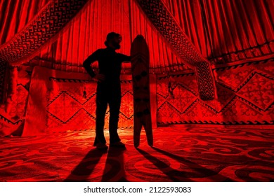 Silhouette Of Man Holding With Split Snow Board Inside Yurt Nomadic House Against Carpet With Ethnic Patterns Lit By Red Light Interior. Backcountry Rider Sport Concept.