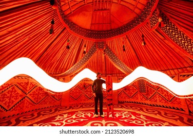 Silhouette Of Man Holding With Split Snow Board Inside Yurt Nomadic House Against Carpet With Ethnic Patterns Lit By Red Light Interior. Backcountry Rider Sport Concept.