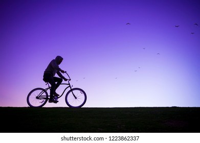 Silhouette Of Man In Hoodie Riding A Beach Cruiser Bicycle Against A Glowing Purple Sky