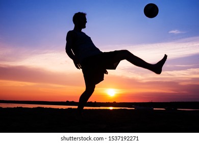 Silhouette of a man juggling a soccer ball on the beach in sunset, surreal blue and orange sky over a sandy beach, sand debris flying around as the man kicks the ball  - Powered by Shutterstock